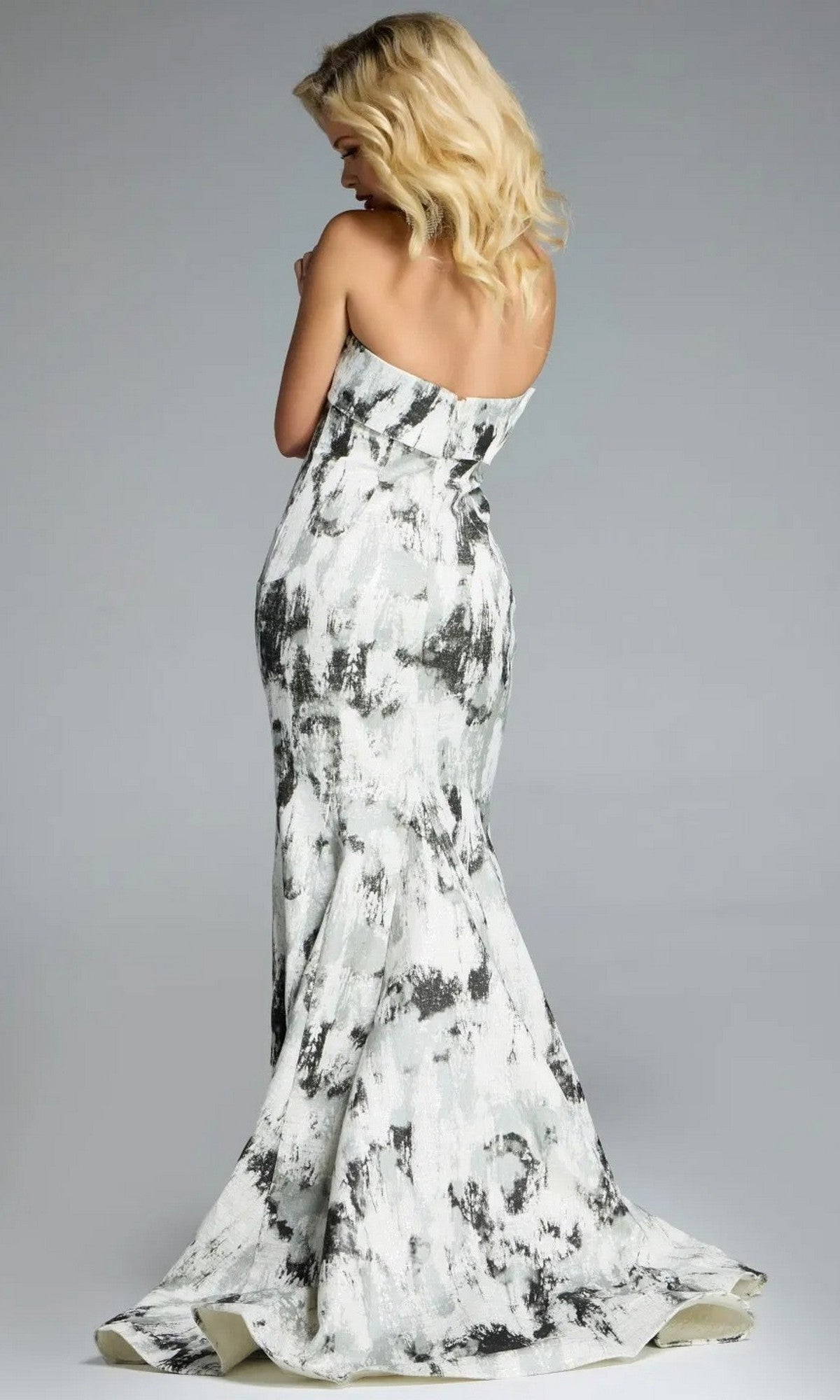 A woman models the Jovani 43977 Long Prom Dress, a strapless, floor-length gown with an abstract print and mermaid silhouette, shown from the back against a plain gray background.